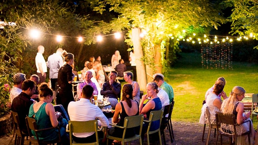 groep jonge mensen zitten 's avonds in kringetjes te praten in de tuin van Vijverschie te praten verlicht door slingers met lampjes in midden-delfland