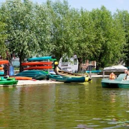 drie mensen in een groene waterfiets met in de achtergrond felgekleurde kano's op de kant voor de verhuur door Kanoverhuur Schipluiden in Midden-Delfland