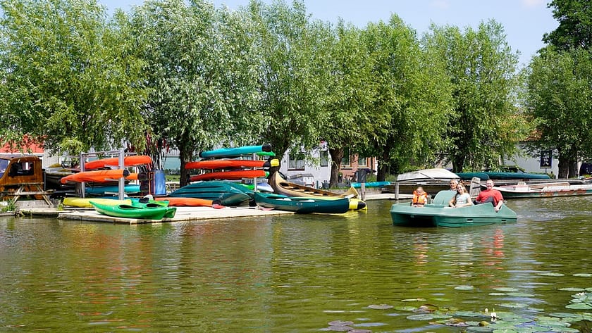 drie mensen in een groene waterfiets met in de achtergrond felgekleurde kano's op de kant voor de verhuur door Kanoverhuur Schipluiden in Midden-Delfland