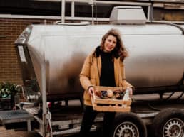 Pauline Schouwenburg De Platte Stad podcast over boeren en de stad Pauline poseert met kratje met streekproducten voor een melktank