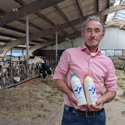 Arnold van Adrichem van Delflandshof in een stal met koeien en twee flessen zuivel van Delflandshof in zijn handen yoghurt melk maker in midden-delfland boer